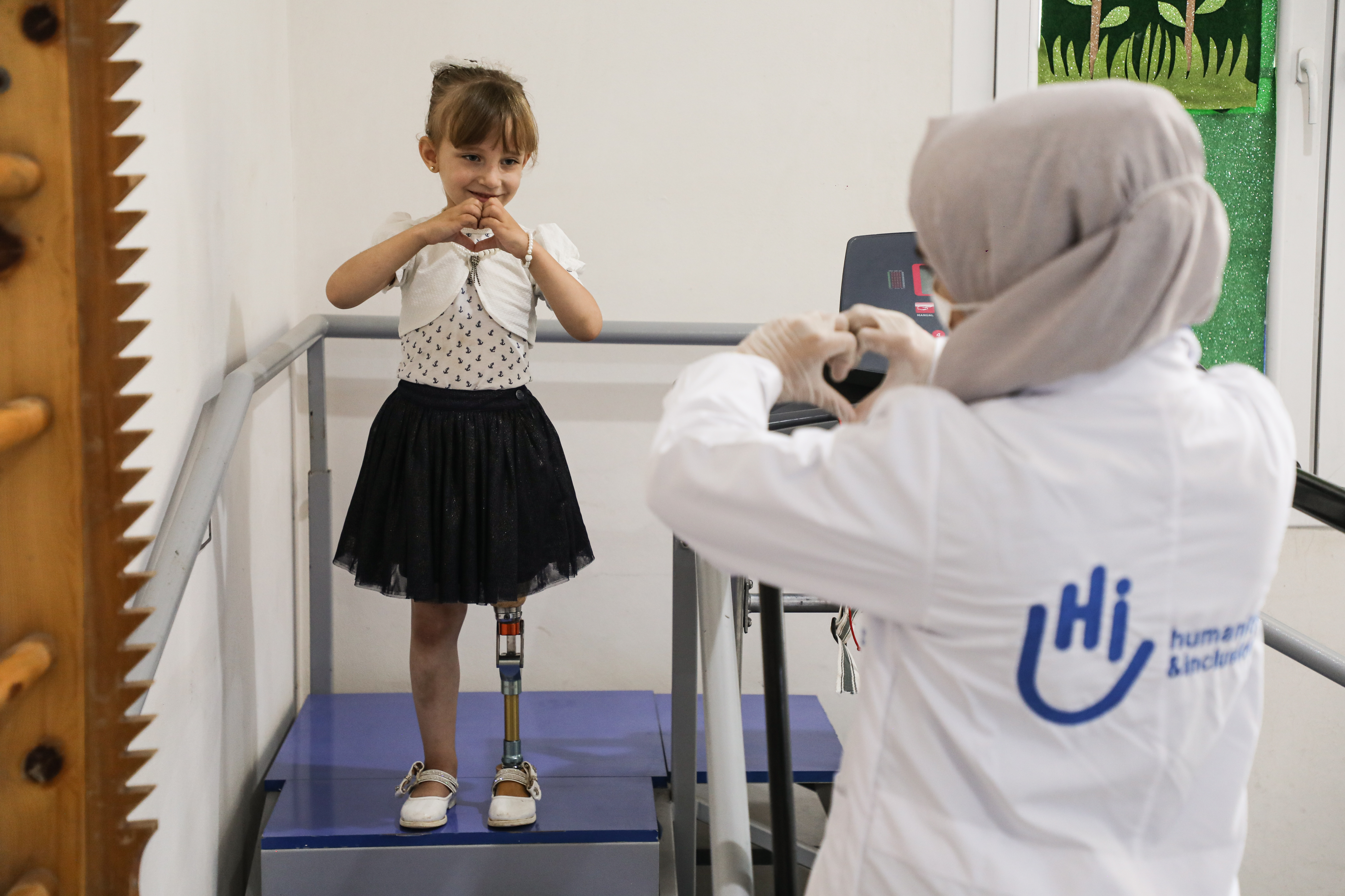 Photo de Enas, une petite fille de 5 ans qui porte une jupe noire avec un haut et un gilet blanc. elle est debout sur un escalier de rééducation avec des rampes. On voit ses jambes, dont sa prothèse de jambe. Enas regarde la travailleuse HI en face d'elle. Enas et Fatima, la travailleuse HI, font un cœur avec leurs mains.