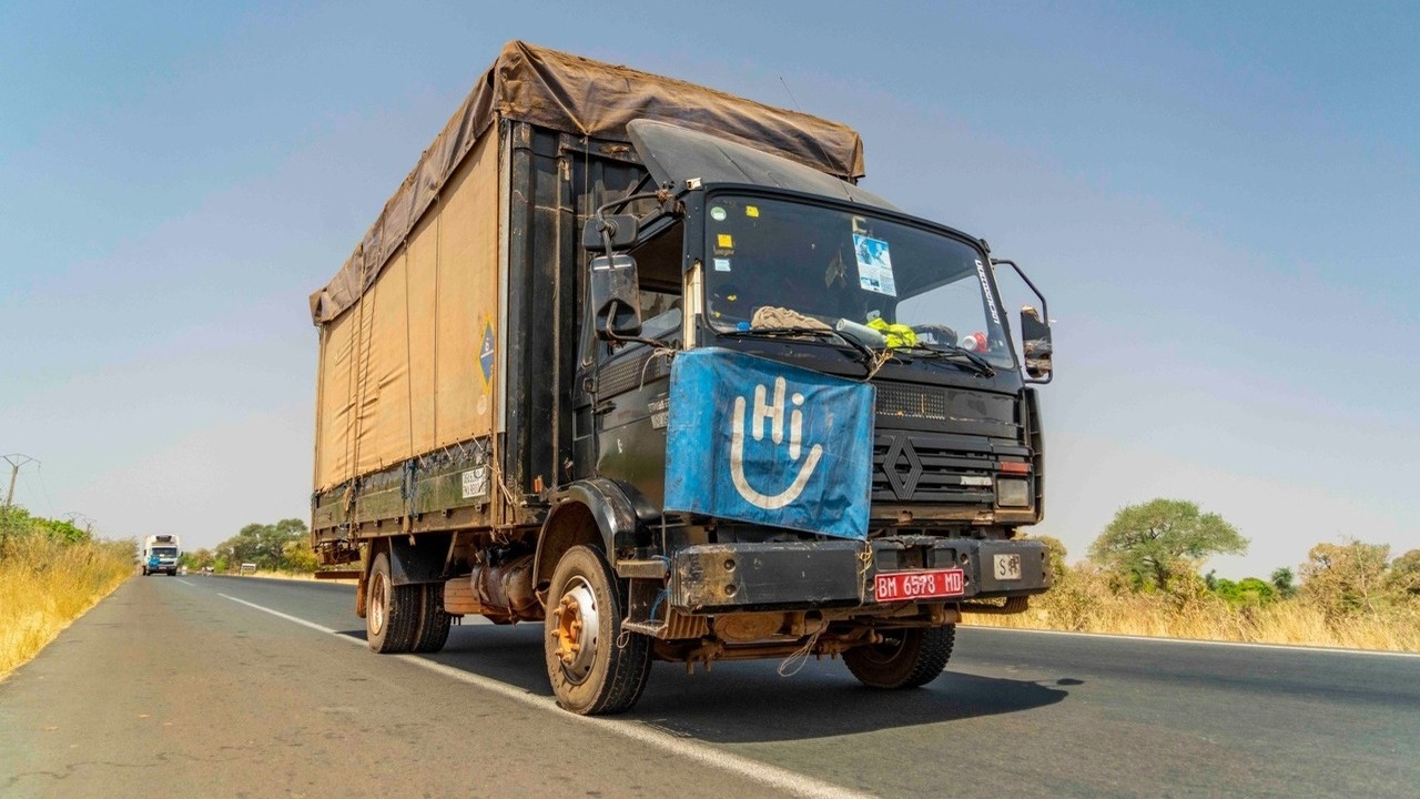 Ein Lastwagen fährt auf einer Straße. Unter der Windschutzscheibe hängt eine blaue Fahne mit dem HI-Logo.