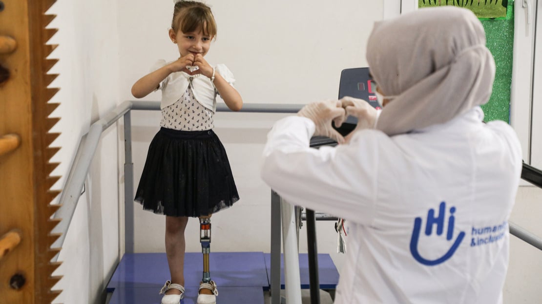 Photo de Enas, une petite fille de 5 ans qui porte une jupe noire avec un haut et un gilet blanc. elle est debout sur un escalier de rééducation avec des rampes. On voit ses jambes, dont sa prothèse de jambe. Enas regarde la travailleuse HI en face d'elle. Enas et Fatima, la travailleuse HI, font un cœur avec leurs mains.