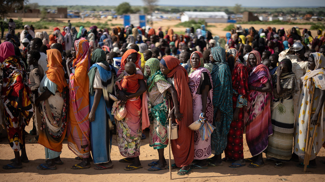 Les femmes se tiennent l'une derrière l'autre, en une ligne que l'on peut voir se poursuivre loin derrière elles. Chacune tient un bassin ou un récipient vide.n a line that can be seen continuing far behind them. Each is holding an empty basin or container.