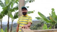 Carlos Bolaños chez lui, dans le département du Cauca. Portrait d'un homme qui s'appuie sur un muret et sourit à la caméra. Derrière lui, on voit des branches de bananiers et on devine au loin des collines verdoyantes.