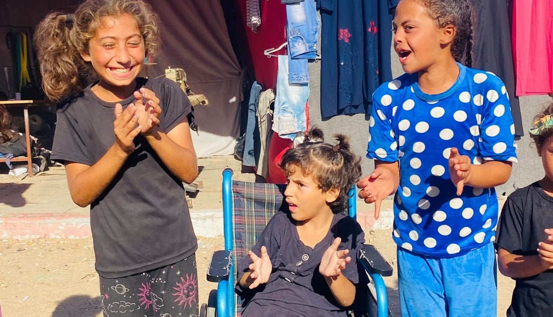 Une petite fille dans son fauteuil roulant bleu entourée de deux autres enfants qui rigolent.